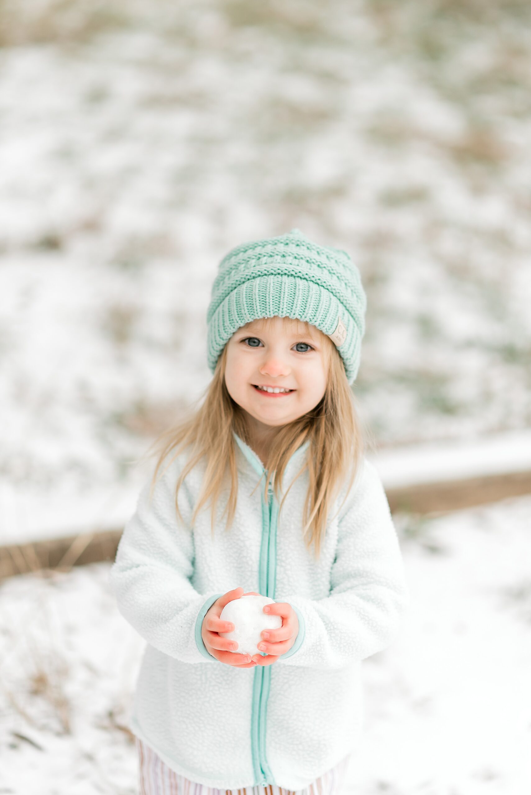 little girl in teal hat holds snowball