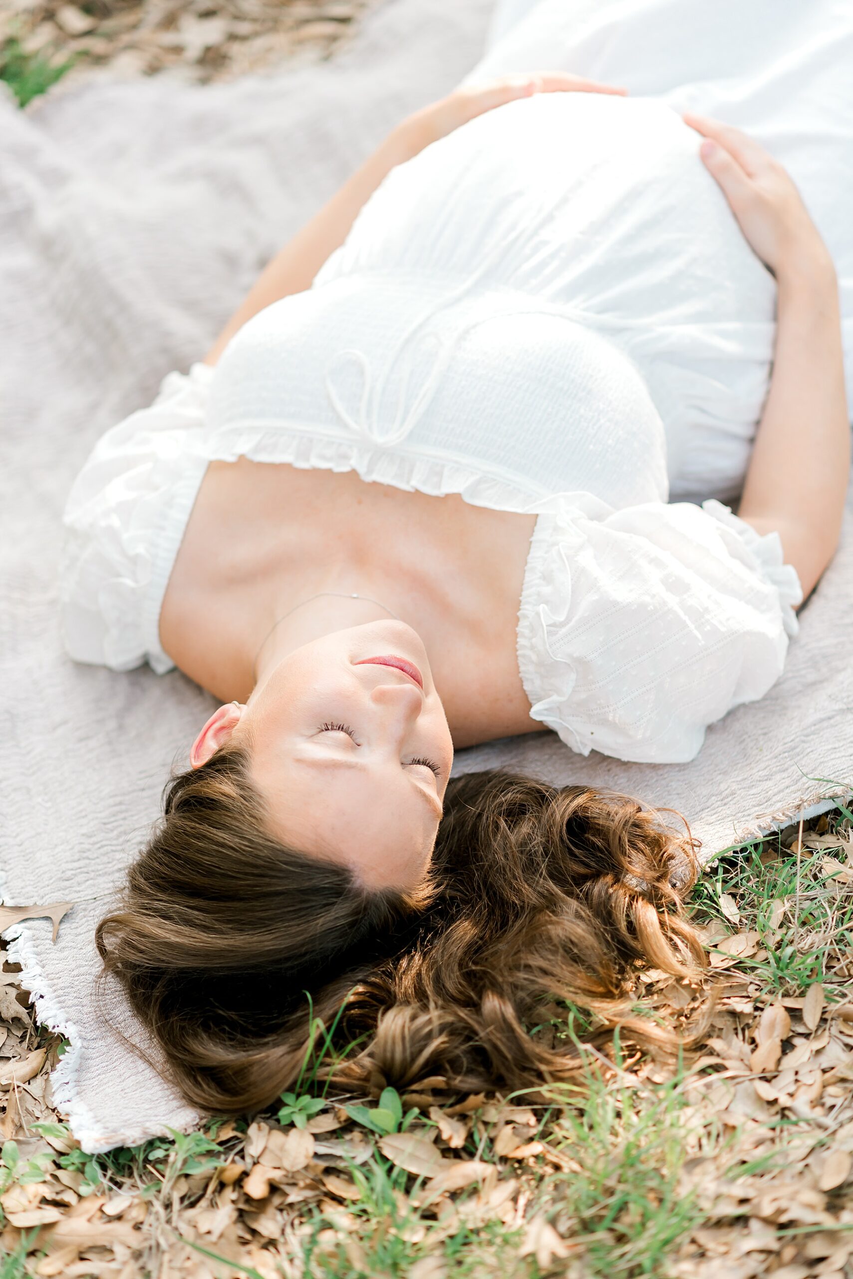 expectant mom in white dress lays on blanket with hands around baby bump