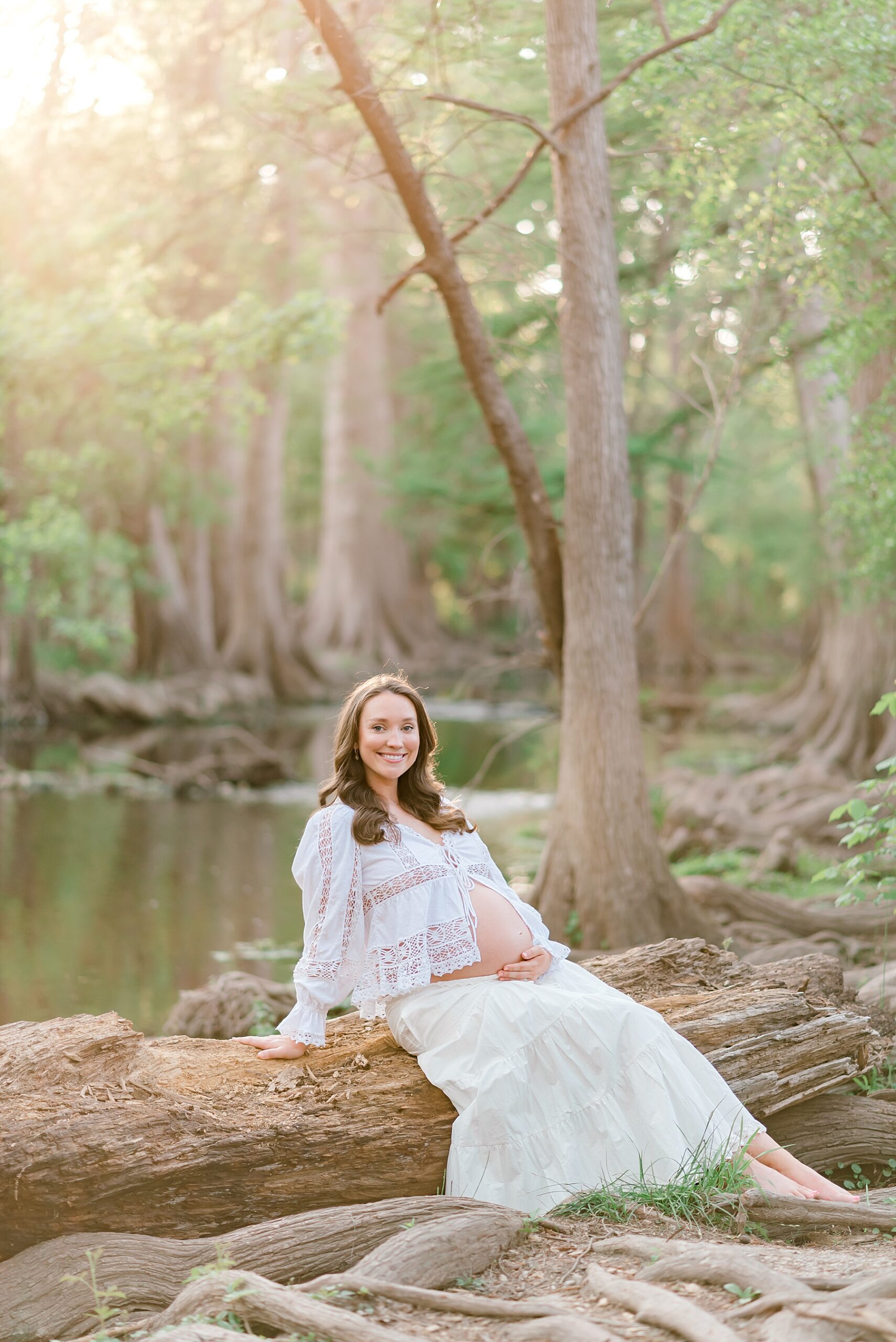 expectant mom sits on large tree log in white skirt and open shirt to show off baby bump