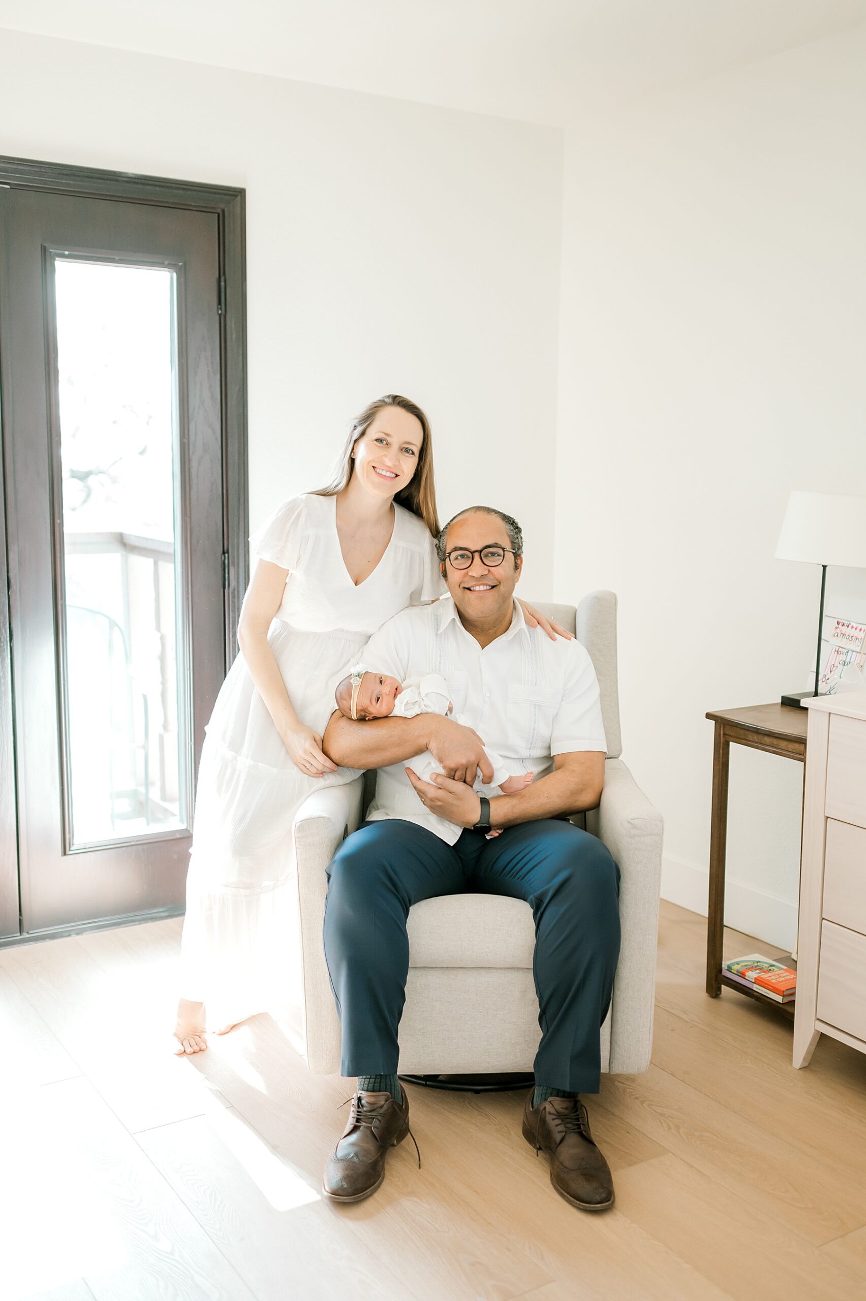 dad sits in chair holding newborn girl with mom next to them