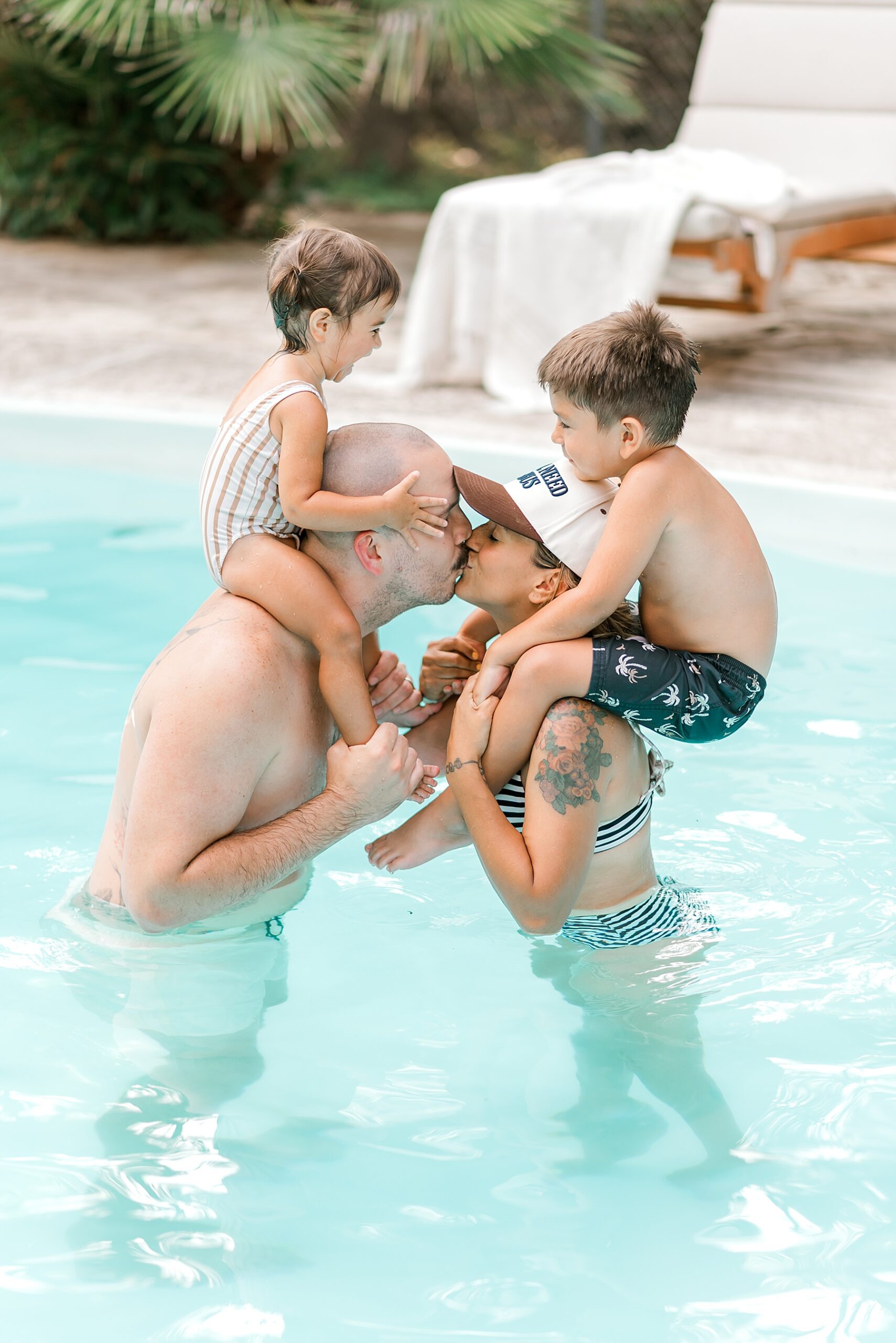 parents holding kids on shoulders kiss while standing in pool