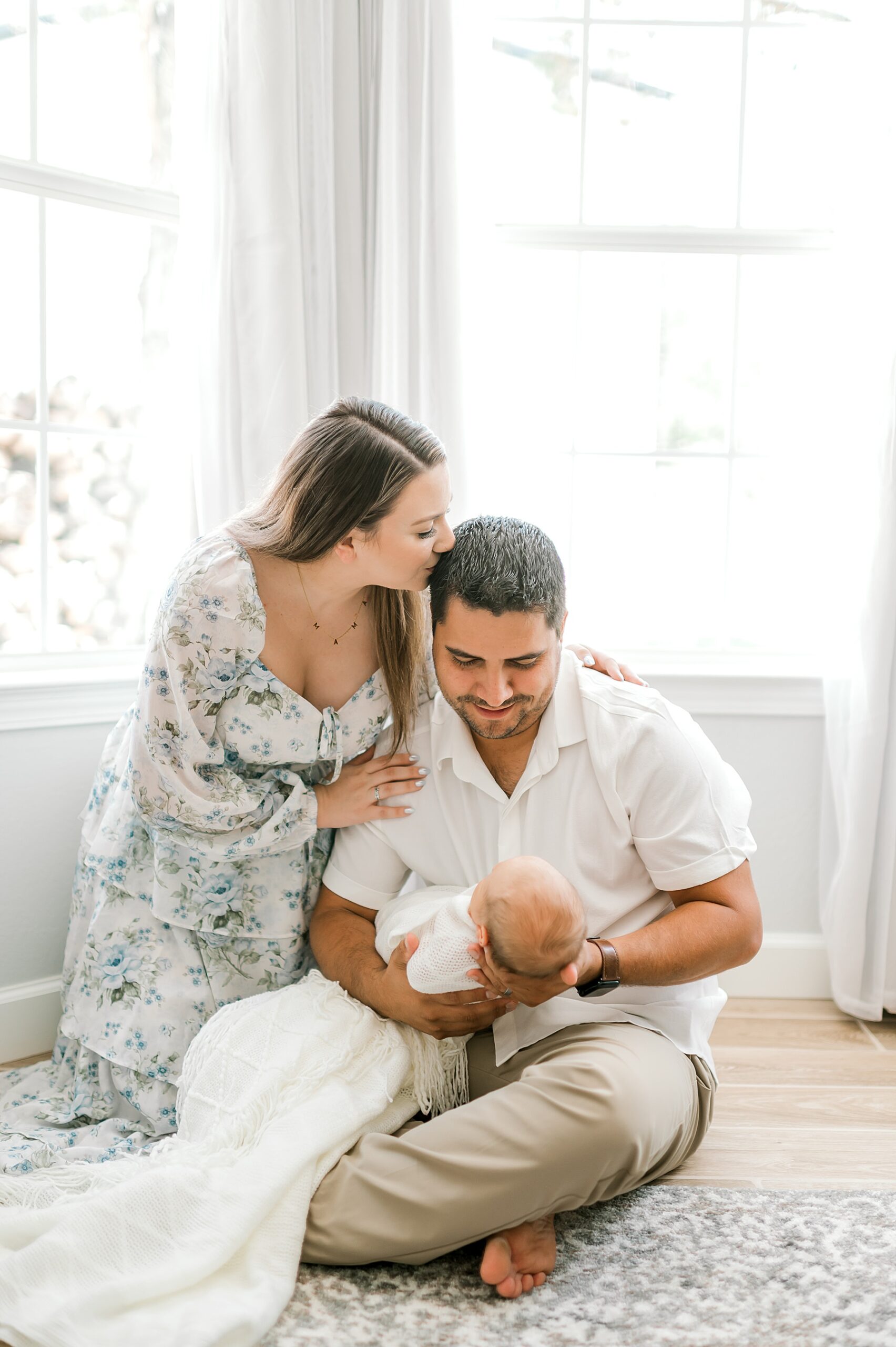 dad sits on floor holding newborn boy as mom kisses her husband's head